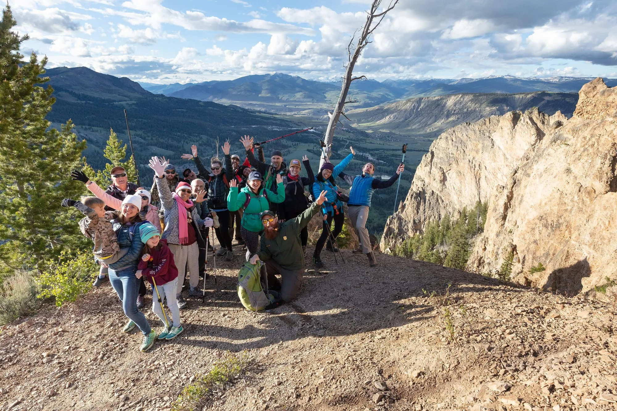Gruppo di escursionisti sorridenti che salutano con le braccia alzate su un sentiero di montagna, luce del tardo pomeriggio